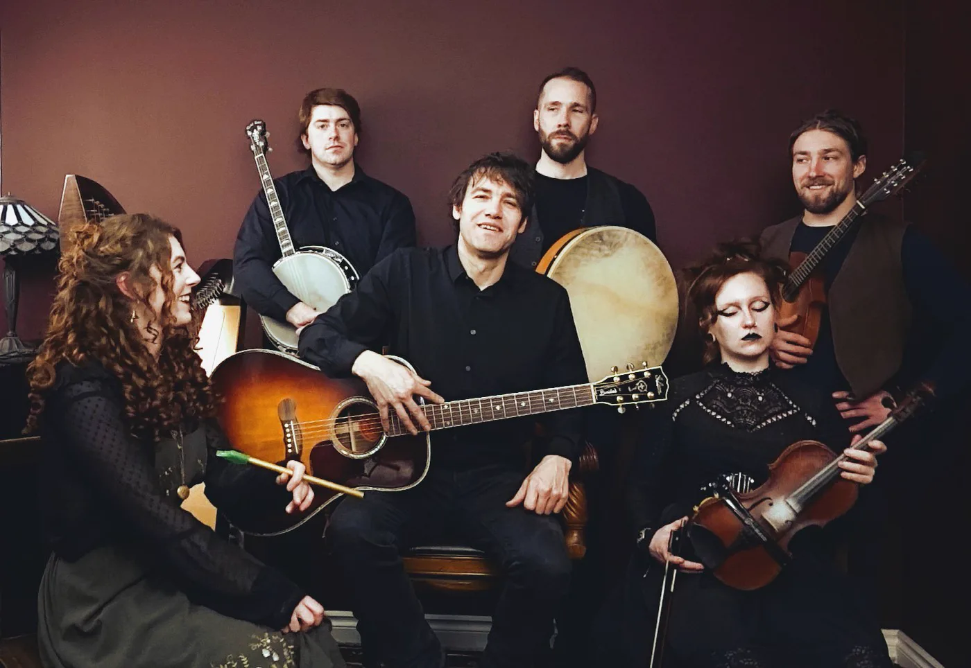 The band as a six piece, seated with guitar, banjo, bodhran, fiddle and mandolin